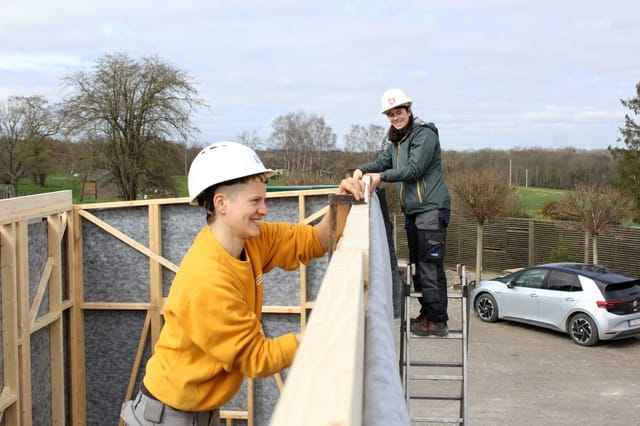 Un participant casqué fixe la lisse haute de l'ossature d'une tiny house