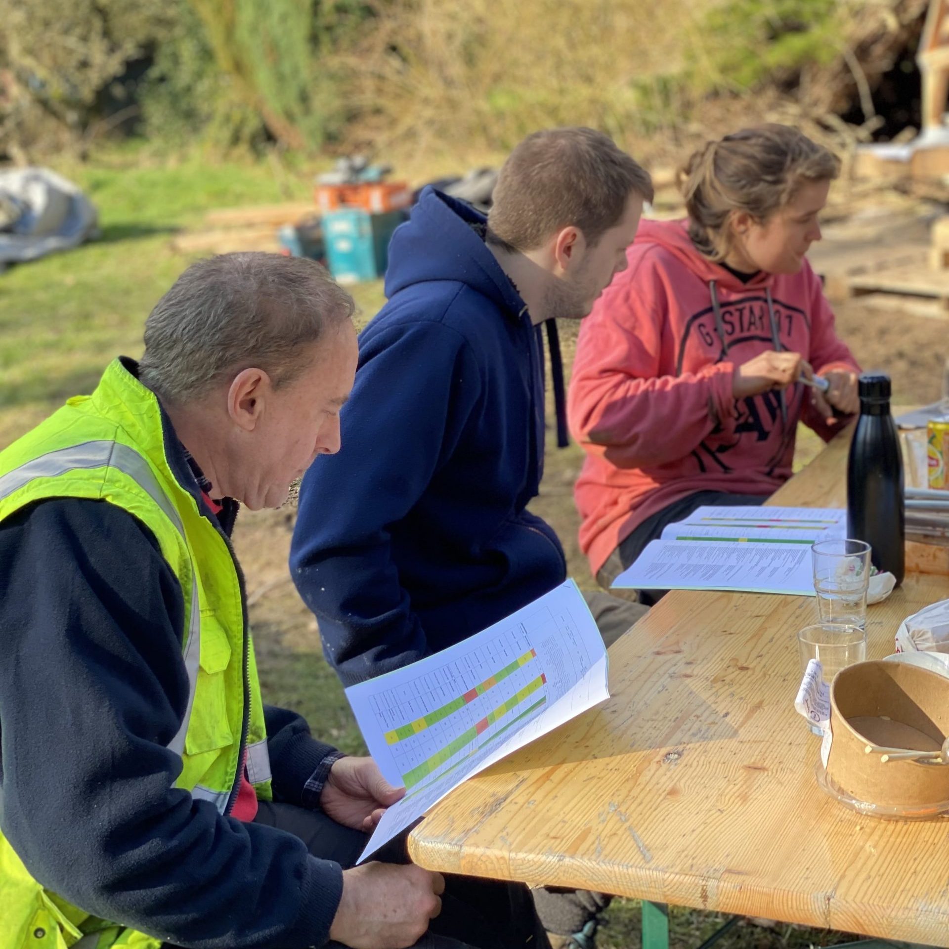 Trois personnes attablées en plein air sur un chantier, penchées sur des plannings imprimés en couleur, gobelets et gourdes posés sur la table en bois.